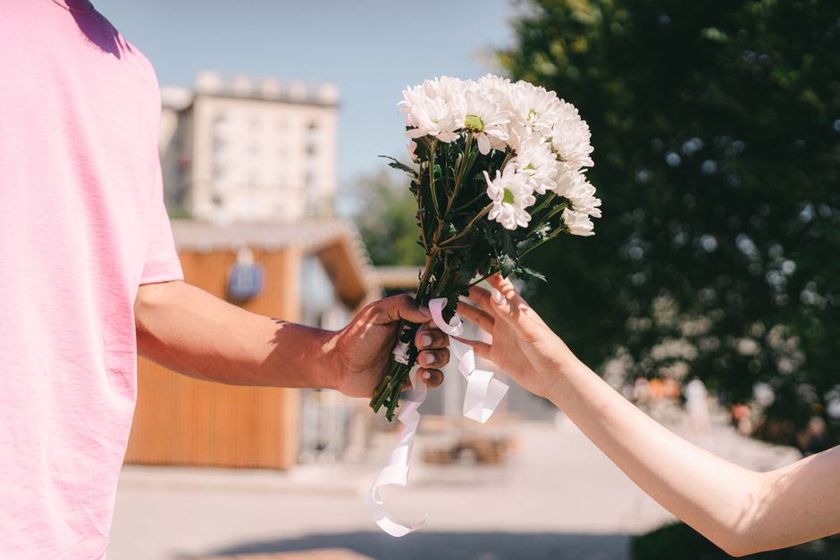 A close-up of a person in a pink shirt handing a floral bouquet to another person outdoors, framed by sunlight and greenery. The bouquet consists of white chrysanthemums and daisies with green stems, 