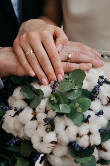 Close-up of a bride and groom's hands with gold wedding bands resting on a bouquet featuring white cotton bolls, sprigs of small blue flowers, and round green eucalyptus leaves. The arrangement is on 